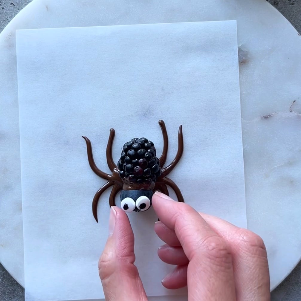 A hand places candy eyes on Chocolate Spiders, a playful treat made of blackberries and chocolate, arranged on parchment paper.