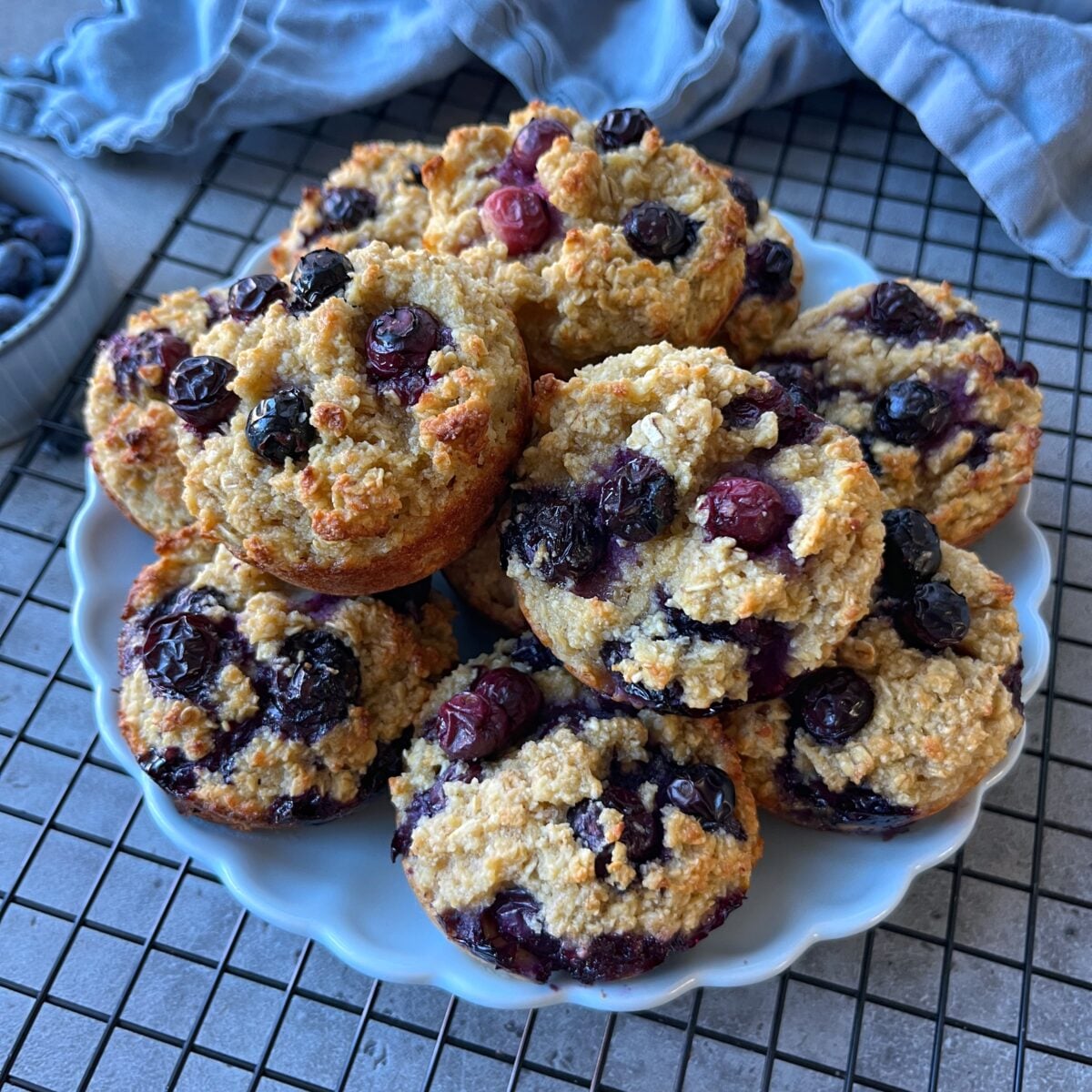 A white plate holds a stack of Easy Breakfast Muffins on a cooling rack, with a bowl of blueberries and a blue cloth visible in the background.