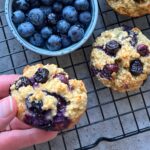 A hand holds a blueberry muffin next to a bowl of fresh blueberries and more muffins, all resting on a black wire cooling rack.