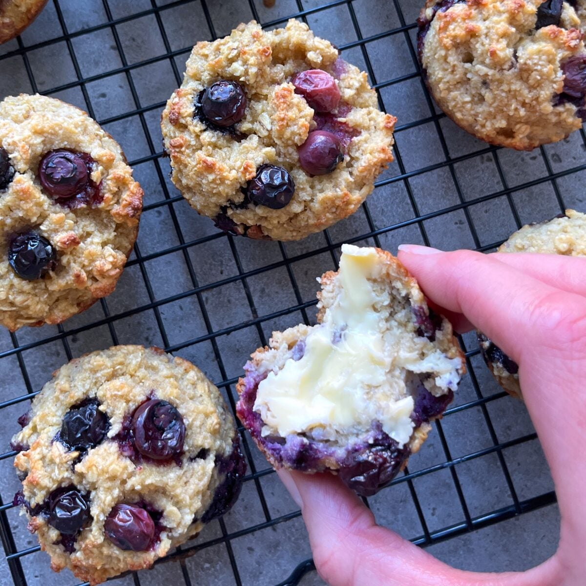 Muffins cooling on a wire rack; a hand holds a halved blueberry muffin topped with butter.