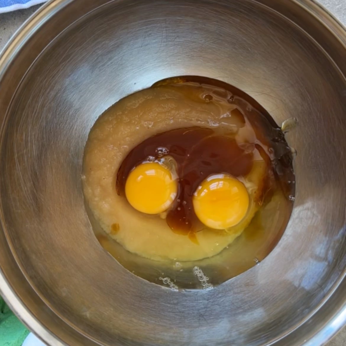 A metal mixing bowl containing two raw eggs, applesauce, and vanilla extract.