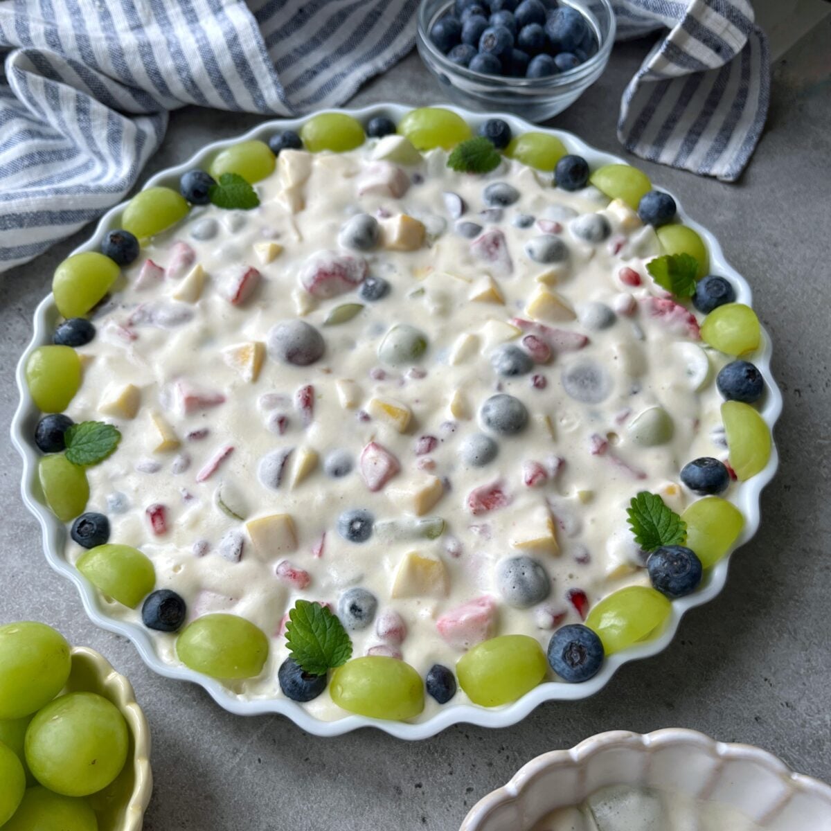 A side view of a round dish filled with creamy fruit salad, topped with a ring of green grapes and scattered blueberries.