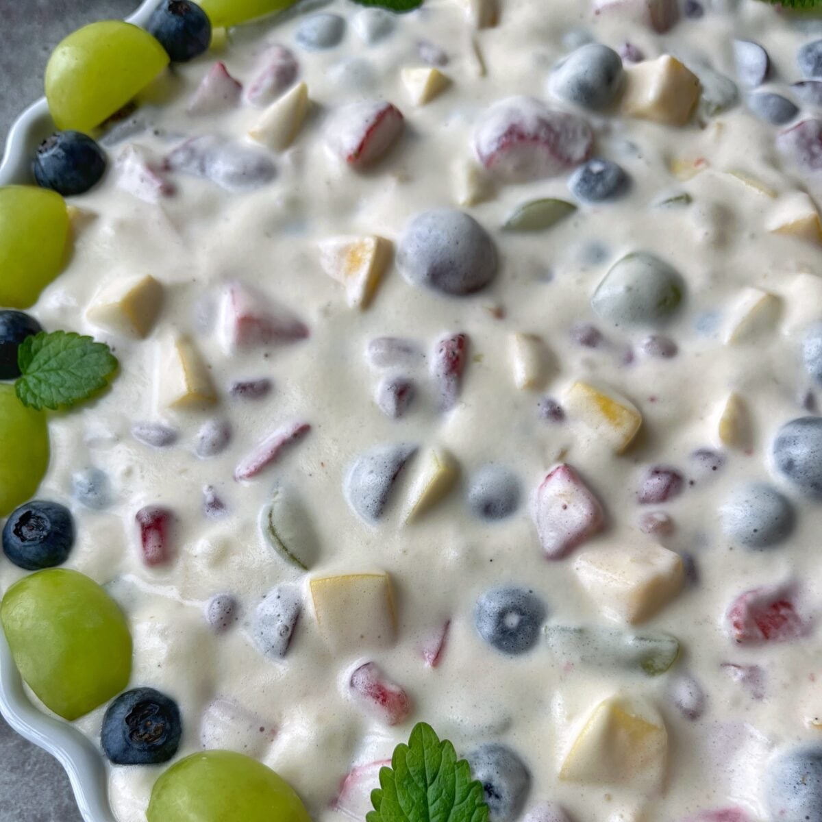 A close-up of a creamy fruit salad with visible chunks of strawberries, grapes, and mango, garnished with green grapes, blueberries, and lemon balm leaves.