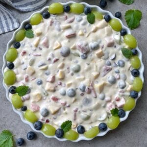 A round dish filled with creamy fruit salad, topped with green grapes, blueberries, and lemon balm leaves, is placed on a gray surface next to a striped napkin.