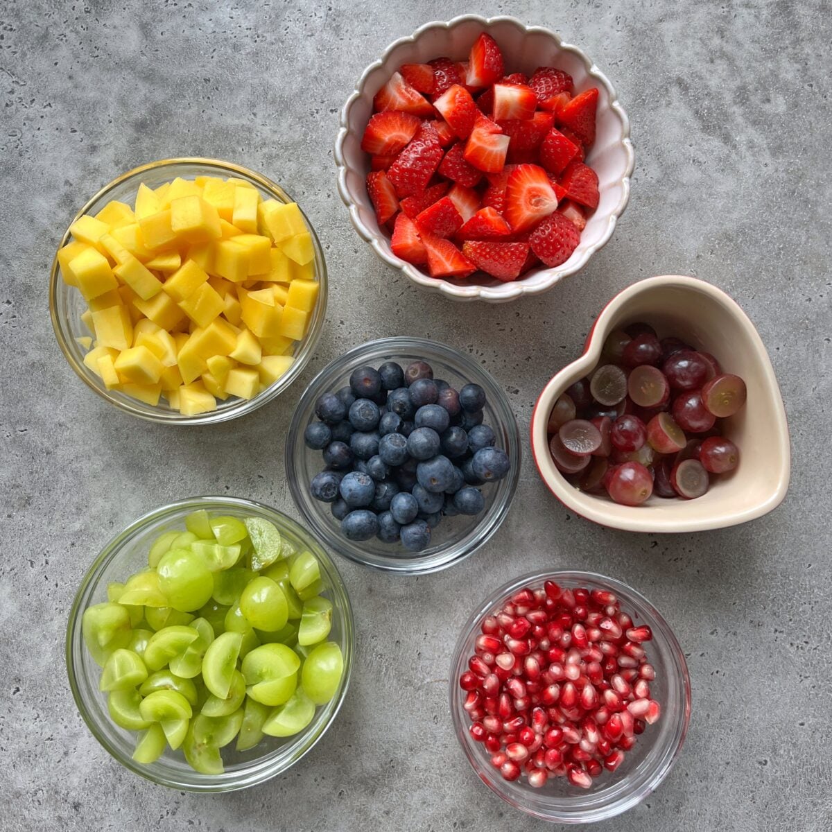 Six bowls of cut fruit: mango, strawberries, red grapes, green grapes, blueberries, and pomegranate seeds.