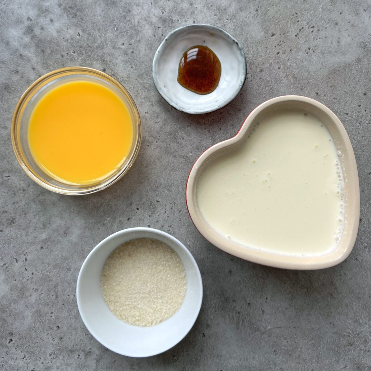 Four ingredients on a gray surface: egg yolks, vanilla paste, whipping cream, and cane sugar.