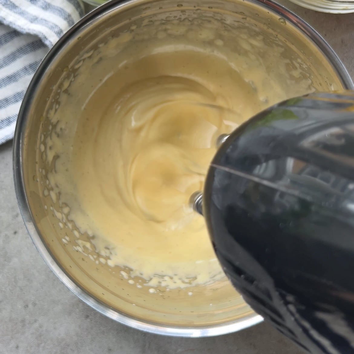 A handheld electric mixer blends a creamy yellow custard mixture in a stainless steel bowl.