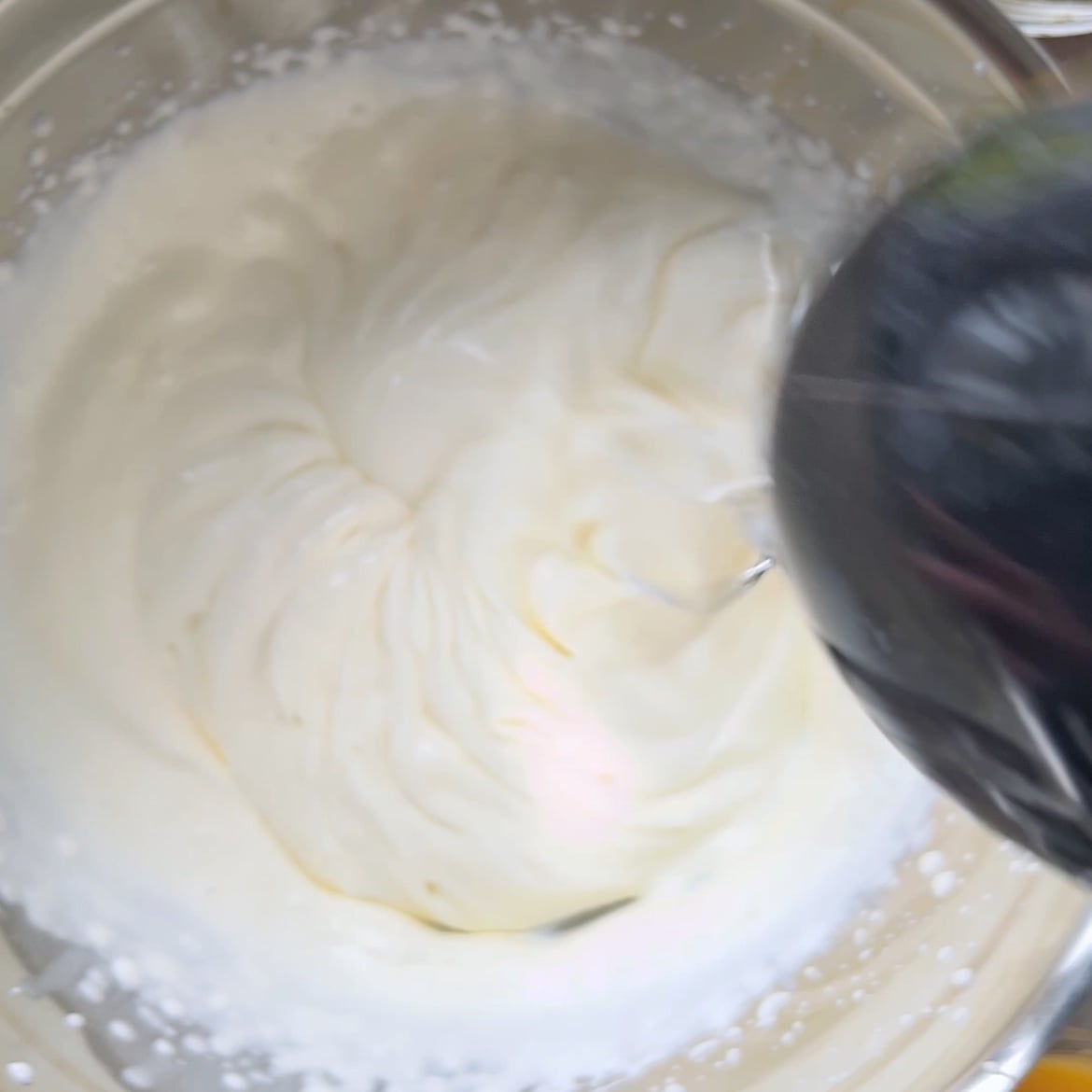 An electric mixer is whipping cream in a metal bowl, creating thick, smooth peaks.