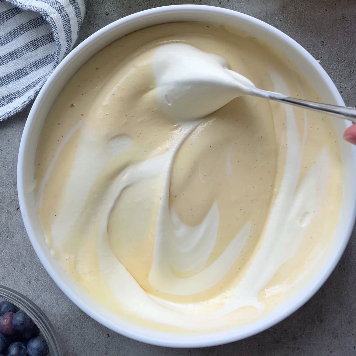 A bowl of pale yellow custard is being mixed with a spoon, partially swirled with white whipped cream.