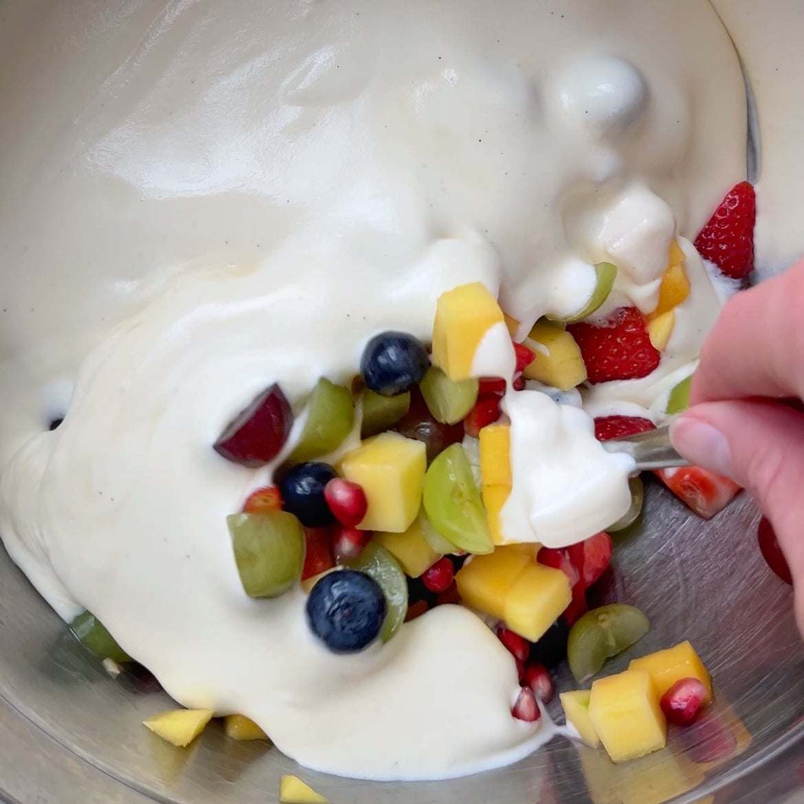 A hand mixes fresh fruit pieces with creamy custard in a metal bowl.
