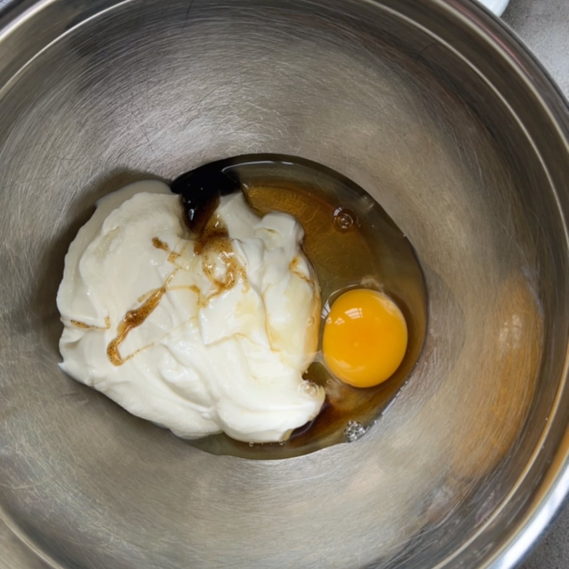 A metal mixing bowl containing Greek yogurt, an egg, and a drizzle of vanilla paste.