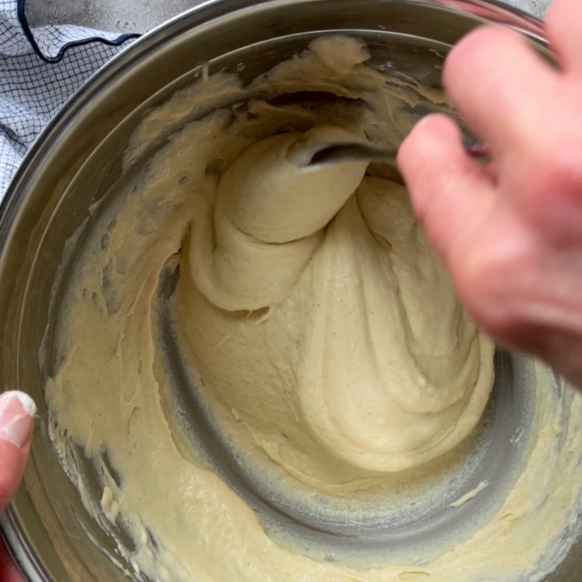 A person stirs thick, pale Greek Yogurt Pancakes batter in a metal mixing bowl with a spoon.