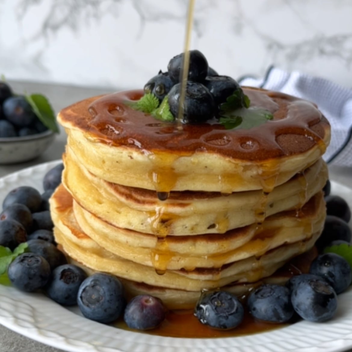 A stack of Greek Yogurt Pancakes topped with blueberries and mint is being drizzled with syrup, with more blueberries scattered around the plate.