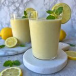 Two glasses of refreshing lemon smoothie, garnished with lemon slices and mint, are displayed on white boards with lemons and mint leaves scattered around.
