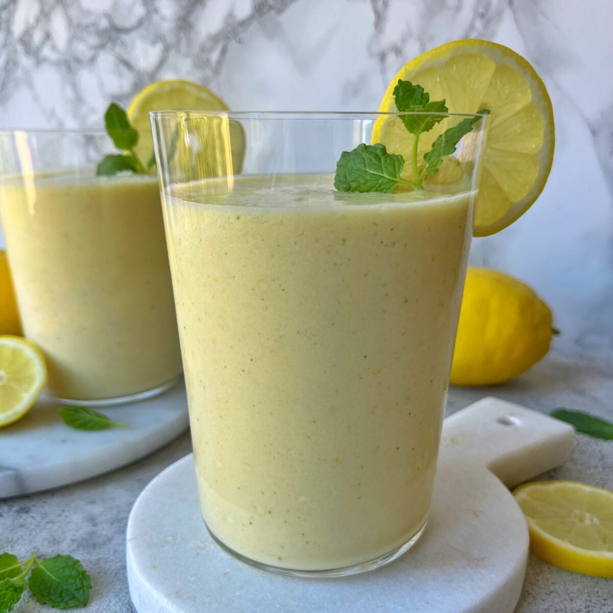 A glass of Lemon Smoothie, yellow and refreshing, is garnished with a lemon slice and mint leaf, while another glass, whole lemons, and fresh mint leaves are in the background.