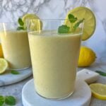 A glass of Lemon Smoothie, yellow and refreshing, is garnished with a lemon slice and mint leaf, while another glass, whole lemons, and fresh mint leaves are in the background.