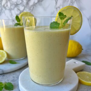 A glass of Lemon Smoothie, yellow and refreshing, is garnished with a lemon slice and mint leaf, while another glass, whole lemons, and fresh mint leaves are in the background.