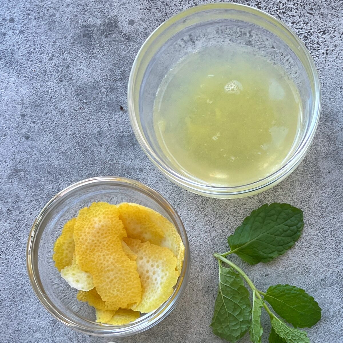 Two glass bowls, one with lemon juice and one with lemon peels, sit on a gray surface next to two sprigs of fresh mint.