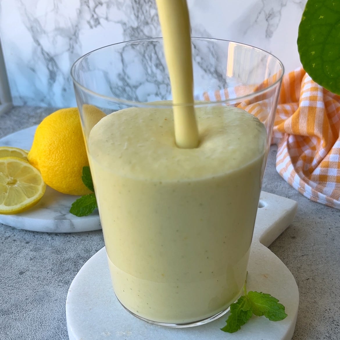 A thick yellow lemon smoothie is being poured into a glass, with fresh lemons, mint leaves, and a yellow checked cloth in the background.