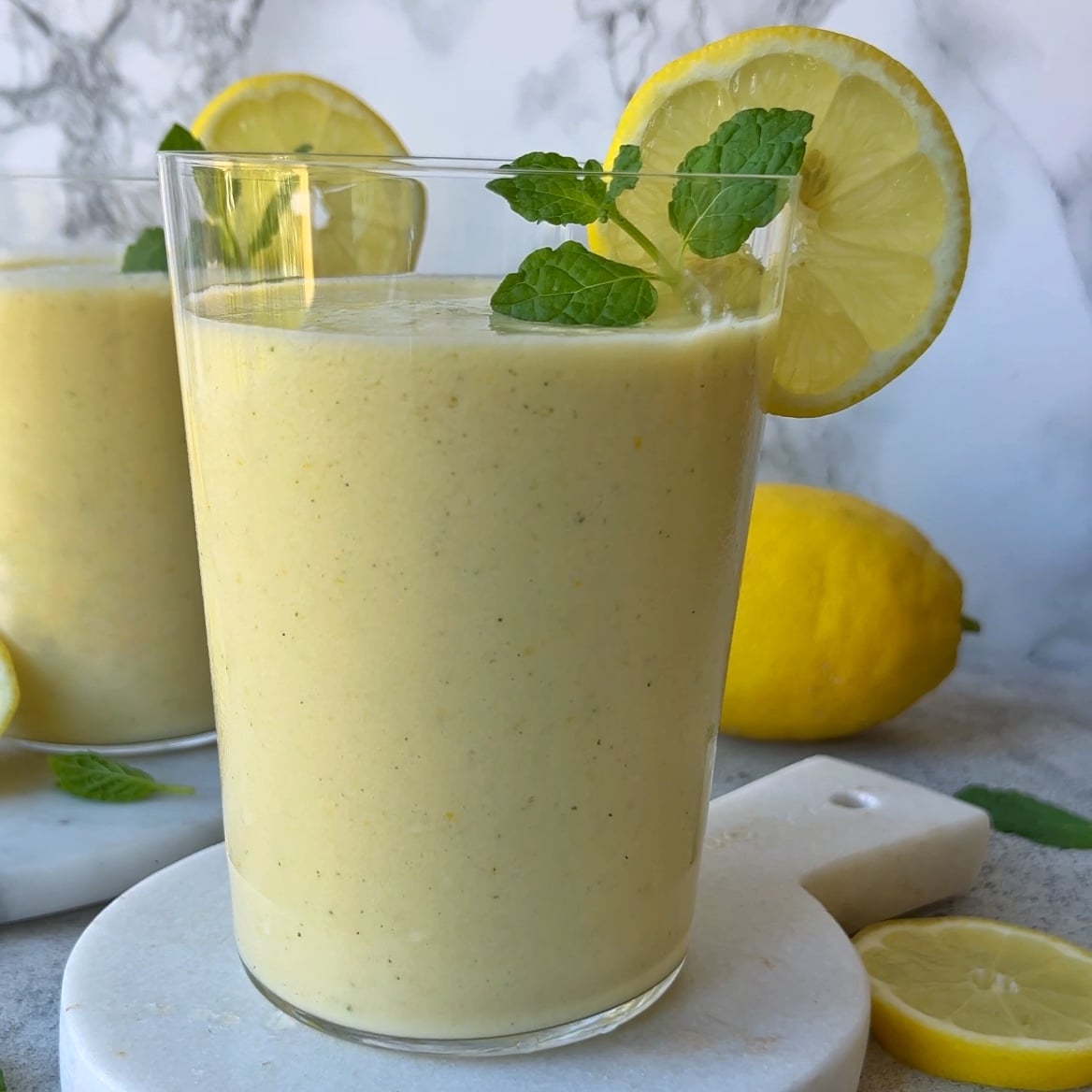 A glass of refreshing Lemon Smoothie garnished with a lemon slice and mint leaves, placed on a white surface with lemons and mint in the background.