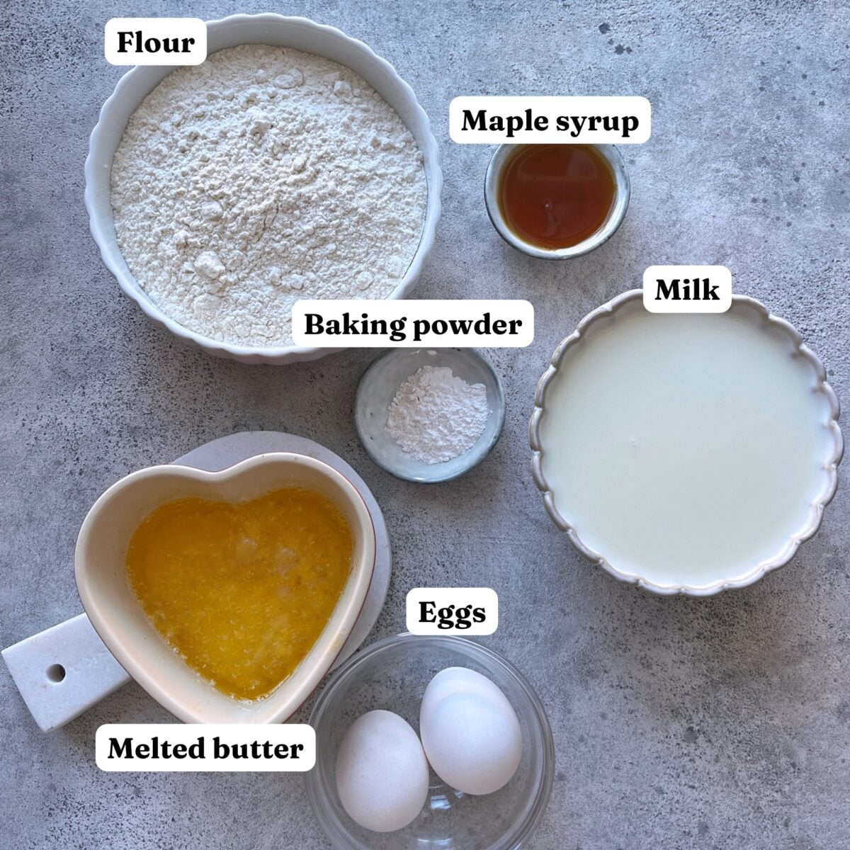 Bowls containing flour, maple syrup, milk, baking powder, melted butter, and two eggs.