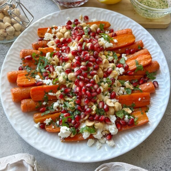 Side view of oven roasted carrots topped with feta, pomegranate, and hasselnuts. A few ingredients are seen in the background.