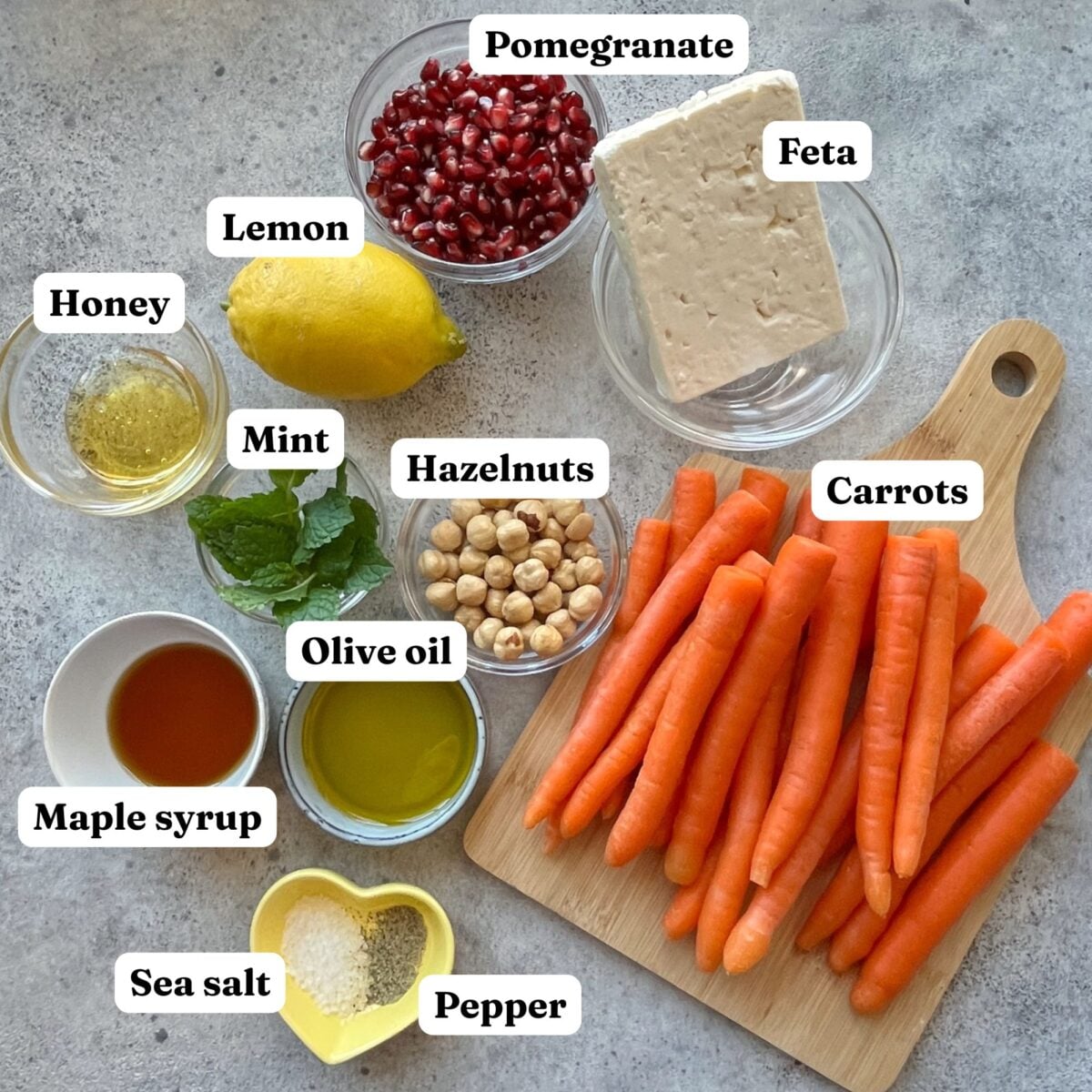 Ingredients for Roasted Maple Carrots are arranged on a surface, including carrots, feta, pomegranate seeds, lemon, honey, mint, hazelnuts, olive oil, maple syrup, sea salt, and pepper.