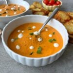 A bowl of creamy tomato soup, garnished with white sour cream dots, black pepper, and fresh basil.