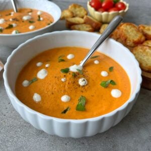 A bowl of creamy tomato soup, garnished with white sour cream dots, black pepper, and fresh basil.