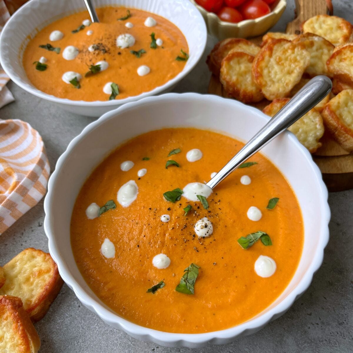 Two bowls of creamy tomato soup, garnished with sour cream dots and herbs, sit with metal spoons, cheese toasts, and a bowl of cherry tomatoes.