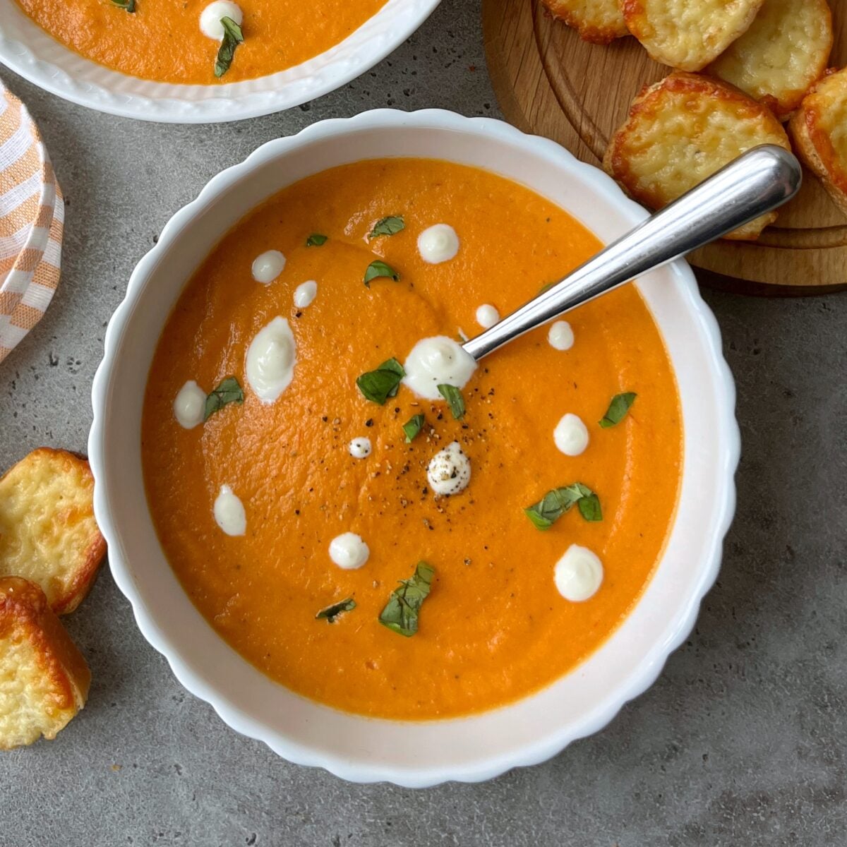 A bowl of creamy tomato soup garnished with white sour cream dots and chopped herbs, served with a spoon. Toasted cheese toasties are off to the side.