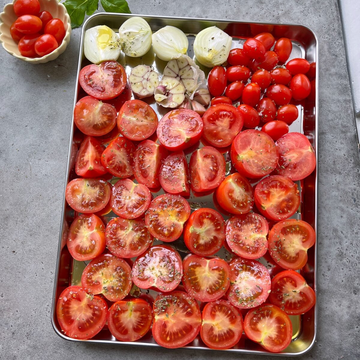 A metal tray with halved tomatoes, onions, a garlic bulb, and grape tomatoes, all seasoned, sits on a gray countertop.