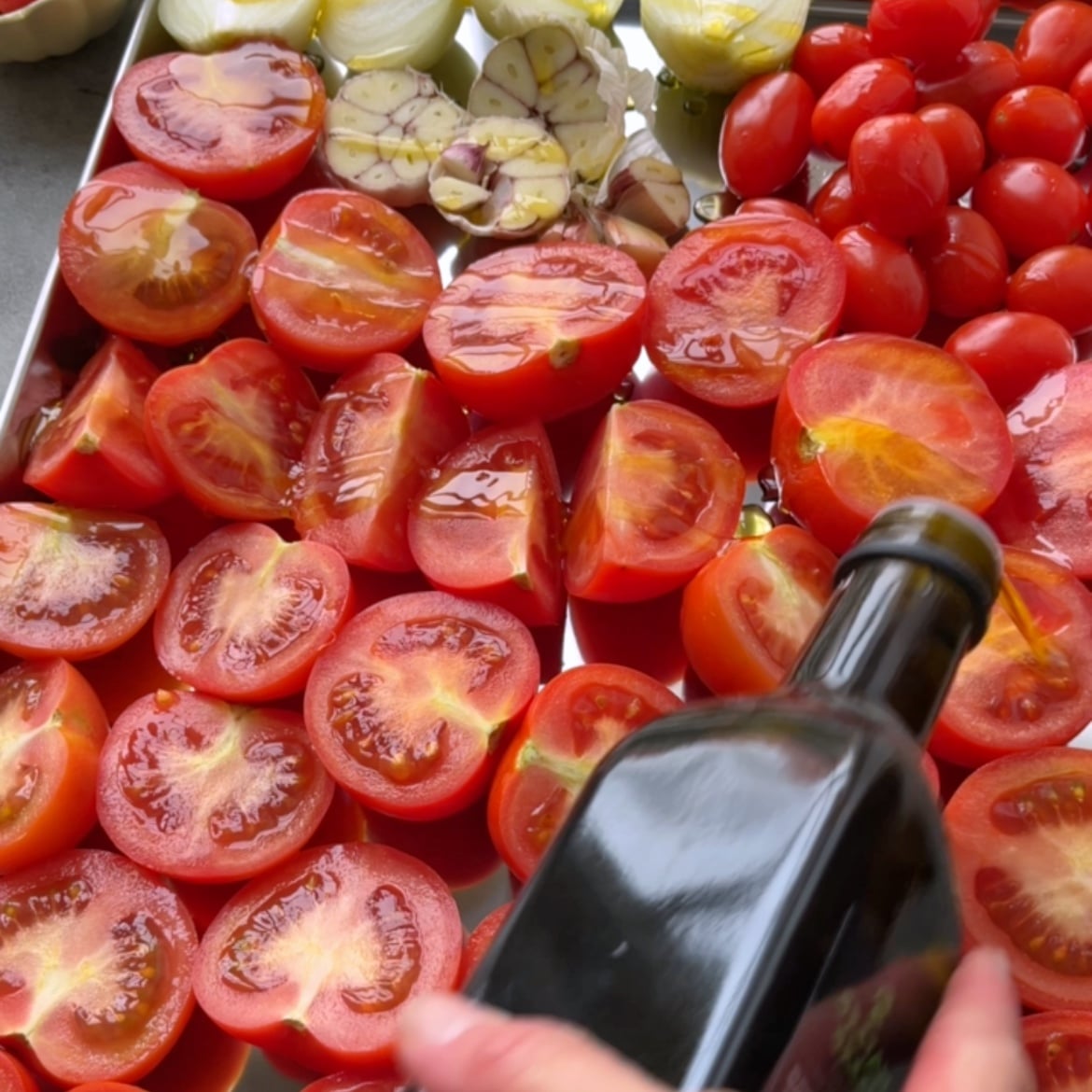 A hand pours olive oil over halved tomatoes, garlic, and onions on a baking tray.