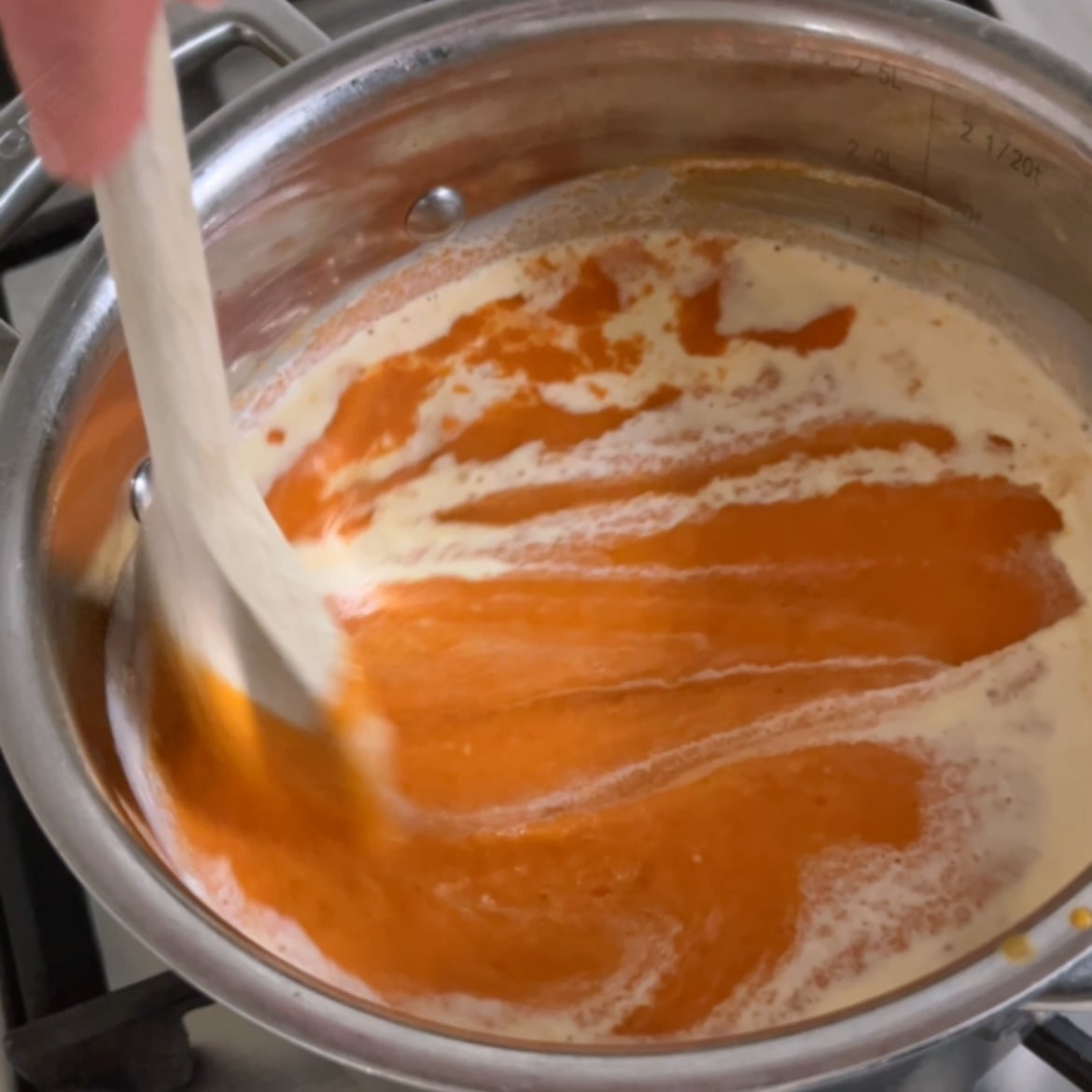 A hand stirs a creamy, orange Roasted Tomato Soup in a stainless steel pot on a stove.