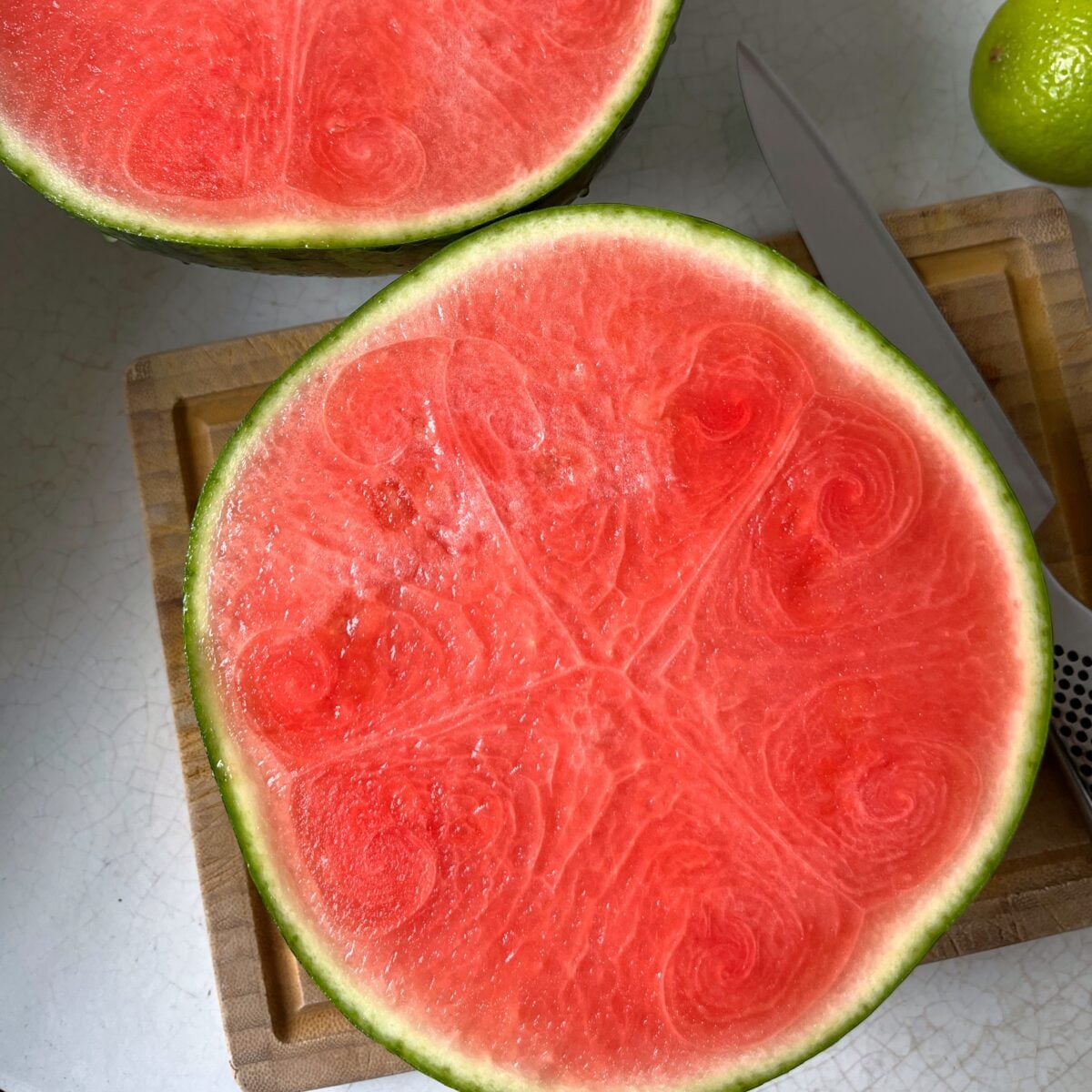 A halved watermelon sits on a wooden cutting board next to a knife and a whole lime.