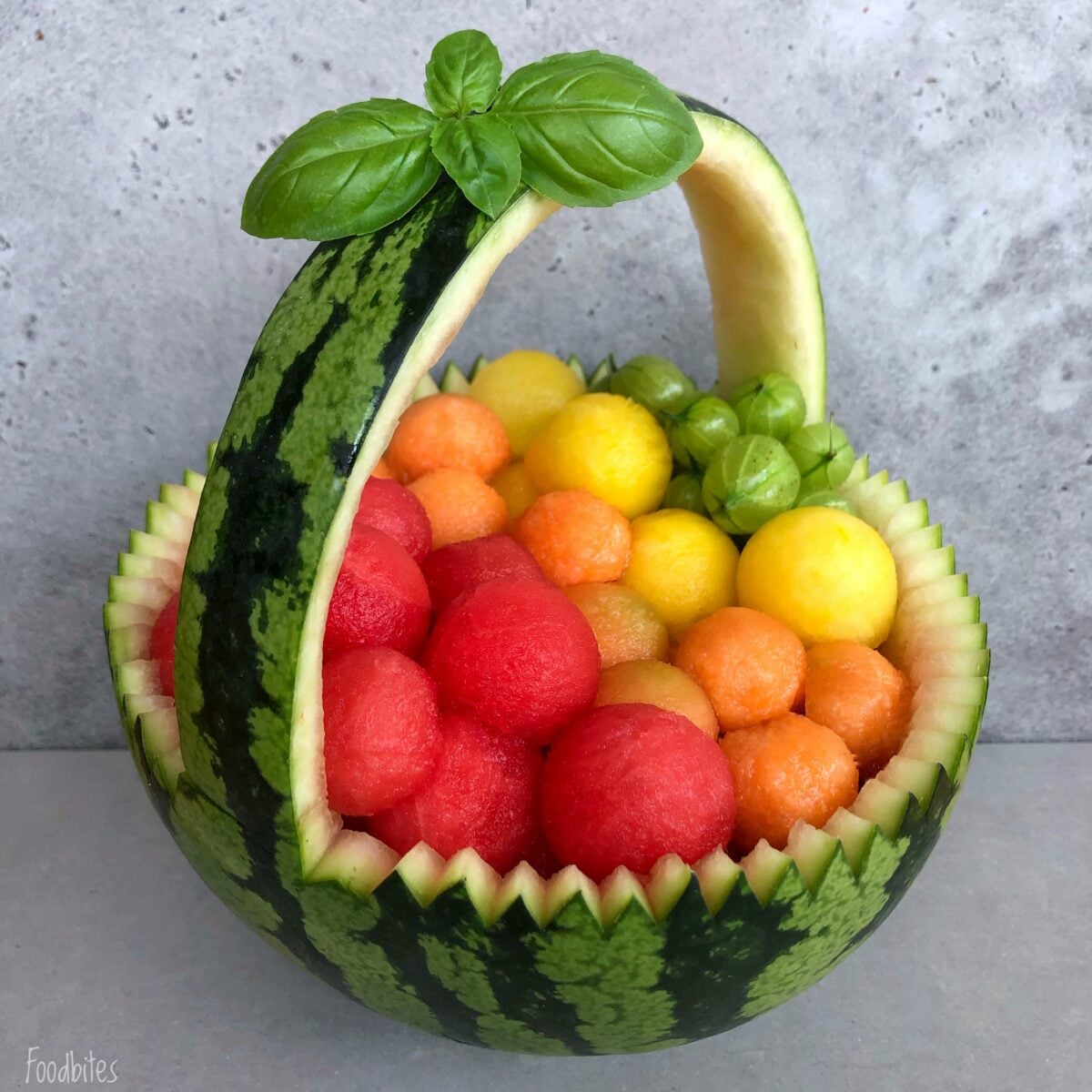A watermelon basket carved into an elegant shape, brimming with colorful melon balls and finished with a garnish of fresh basil.