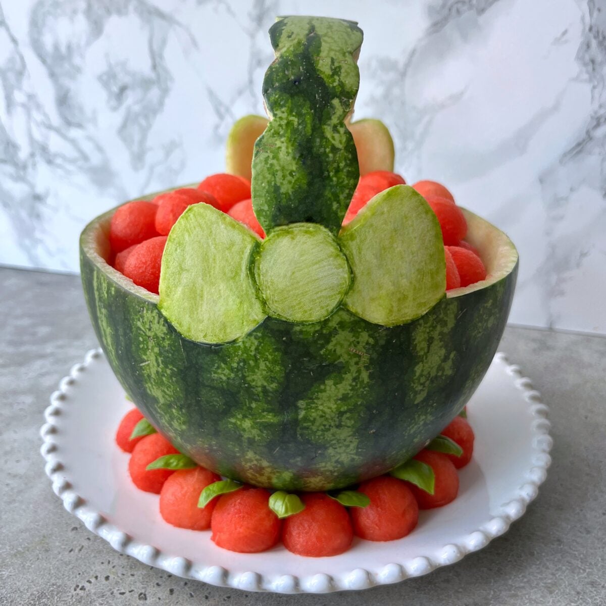 A watermelon basket filled with watermelon balls and decorated with carved green rind bow, sits on a white plate with more watermelon balls arranged around the base.