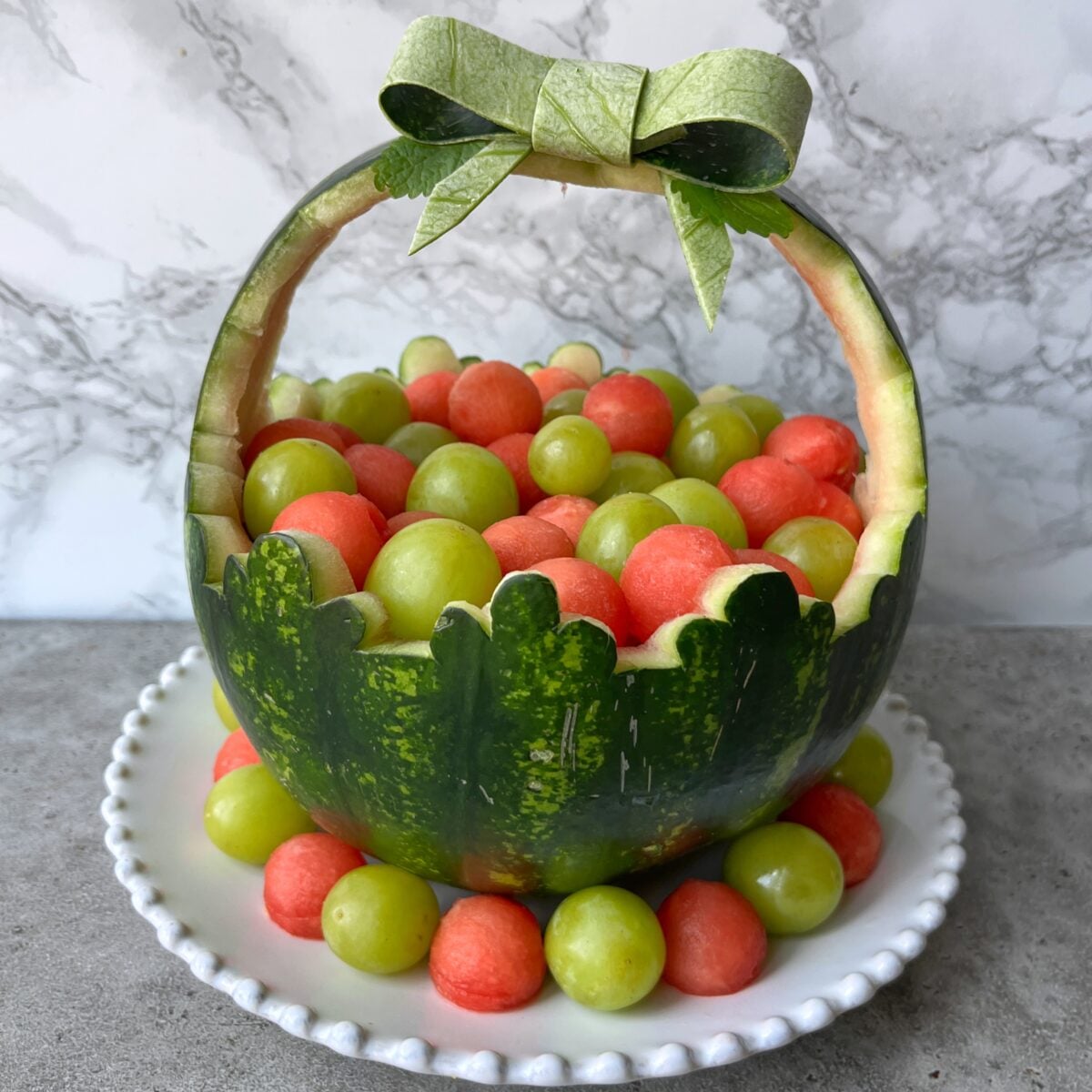 A Watermelon Basket carved into a basket shape, filled with green grapes and watermelon balls, sits on a white plate. The handle is topped with a bow made of watermelon rind.