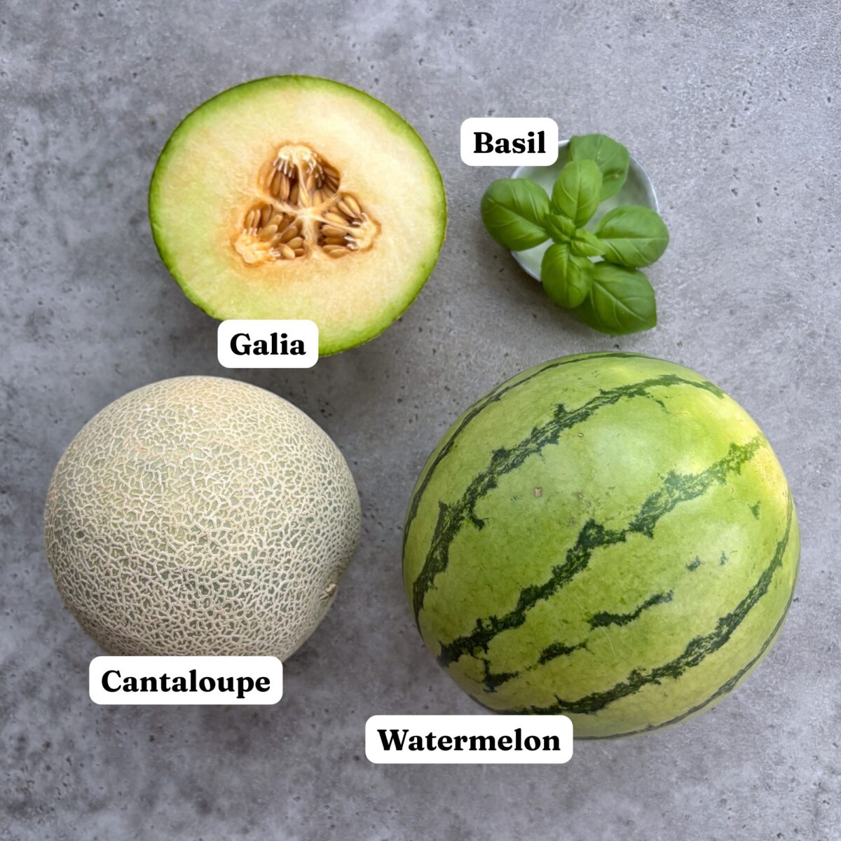 A halved galia melon, a cantaloupe, a watermelon, and fresh basil.