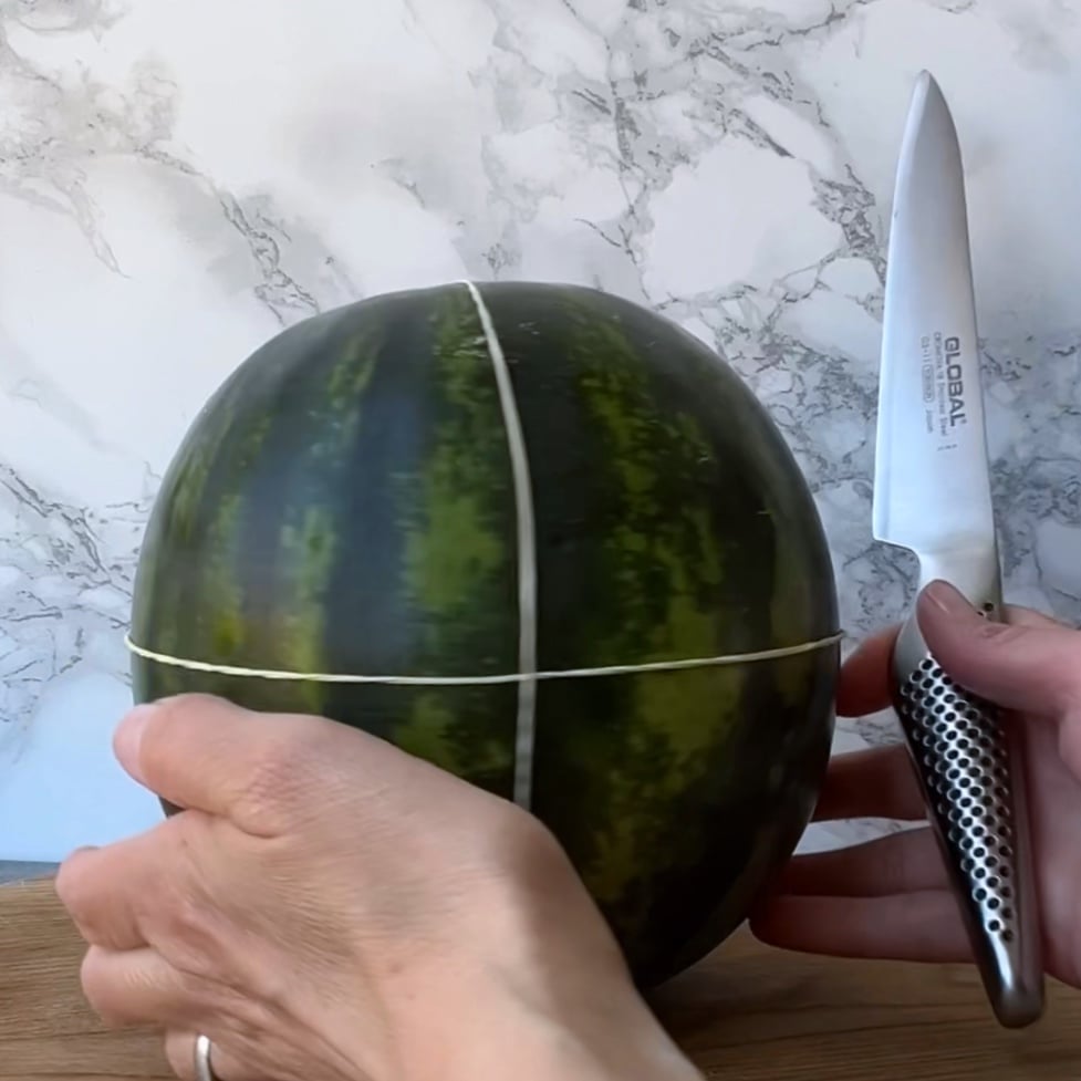 A person holds a knife and a watermelon wrapped with two rubber bands.