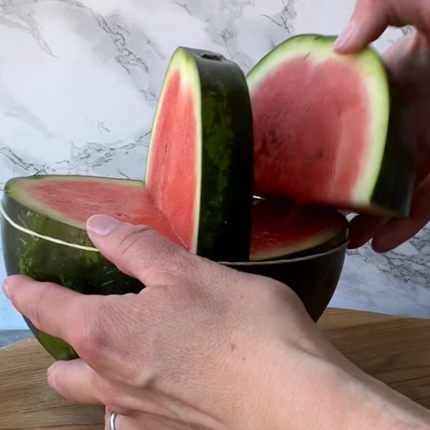A person lifts a large wedge of watermelon from a halved watermelon resting on a wooden surface.