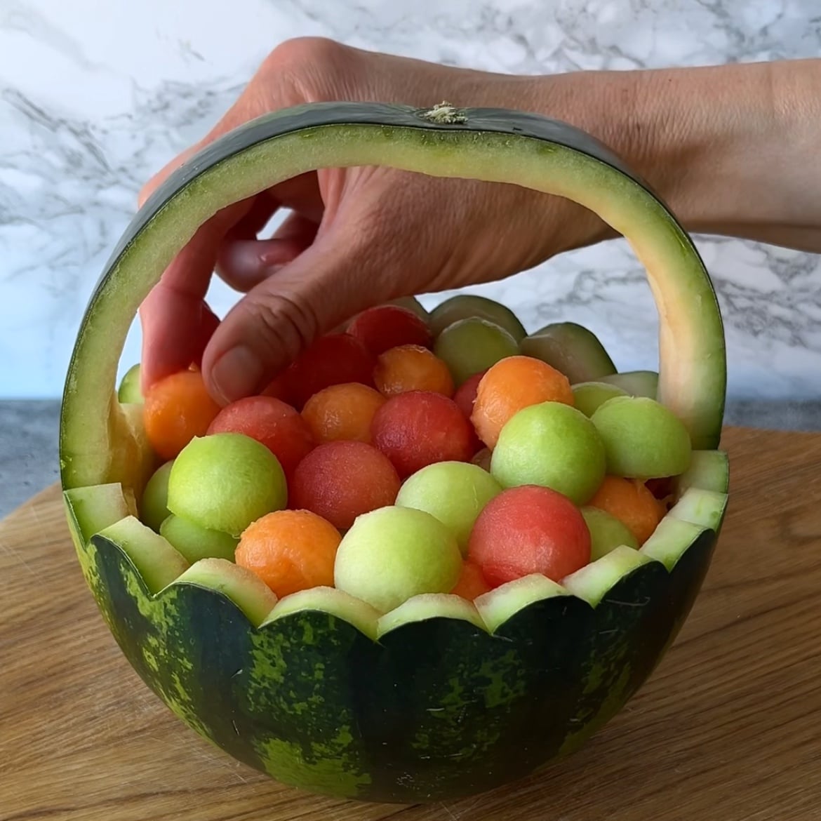 A hand adds assorted melon balls to a watermelon basket.