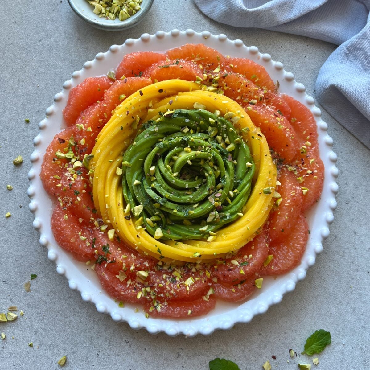 A neatly arranged salad with concentric circles of grapefruit, mango, and avocado slices, topped with chopped pistachios on a white plate.