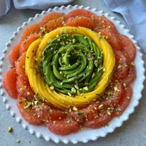 A plate arranged with concentric circles of grapefruit, mango, and avocado slices, topped with chopped pistachios, and a herby dressing.