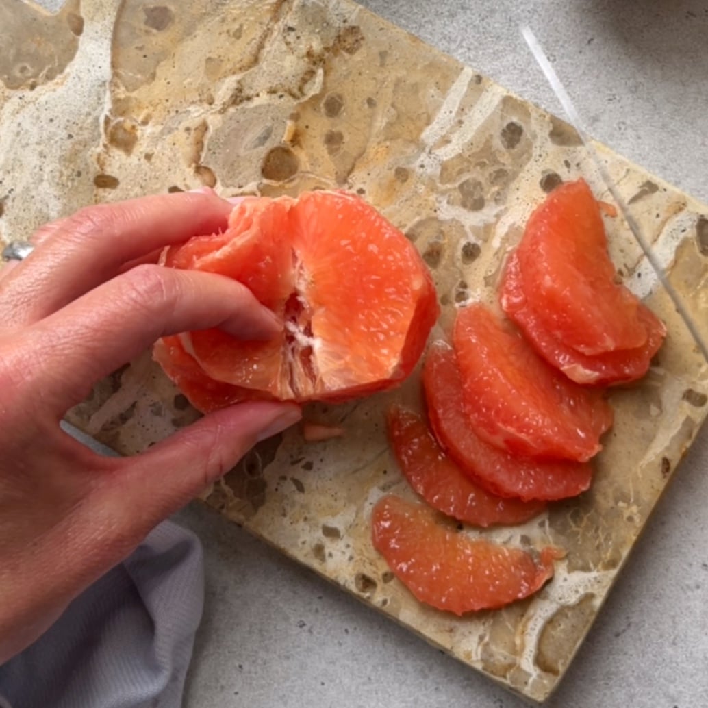 A hand separates grapefruit segments on a marbled cutting board using a knife.