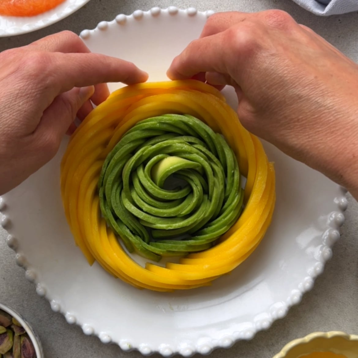 Two hands arrange slices of avocado and mango in a circular pattern on a white plate.