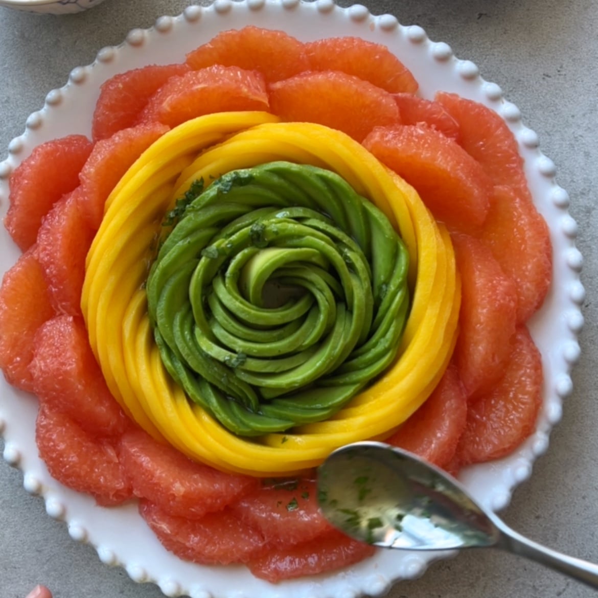 A hand spoons dressing over a salad made with grapefruit slices, mango slices, and avocado slices arranged in a flower pattern.
