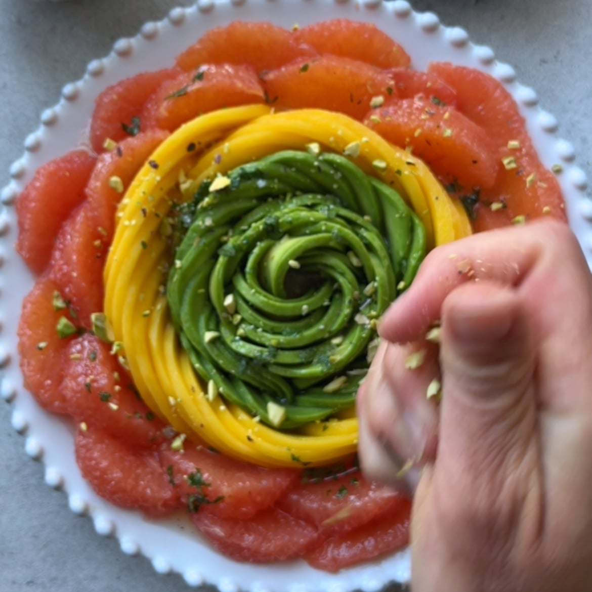 A hand sprinkles chopped nuts over a neatly arranged Avocado Grapefruit Salad.