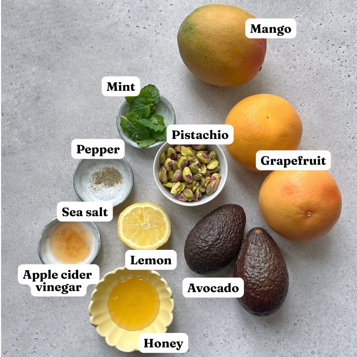 Assorted ingredients on a gray surface: mango, grapefruit, avocado, lemon, honey, apple cider vinegar, sea salt, pepper, mint, and pistachios.