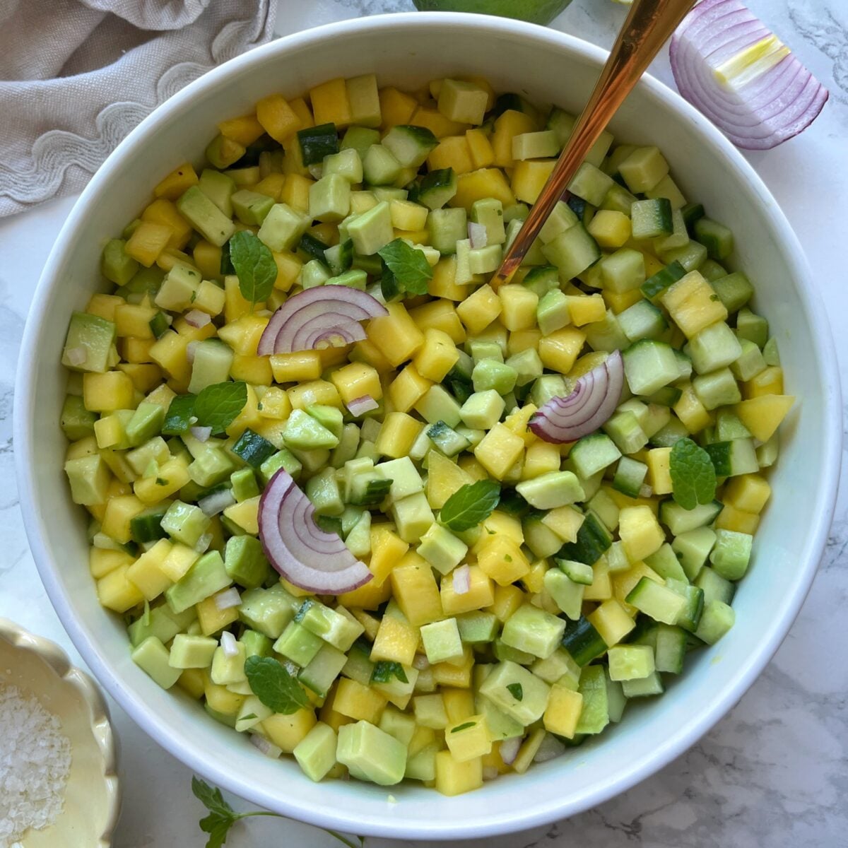 A bowl of diced mango, cucumber, and avocado salad garnished with red onion slices and mint leaves, with a serving spoon.